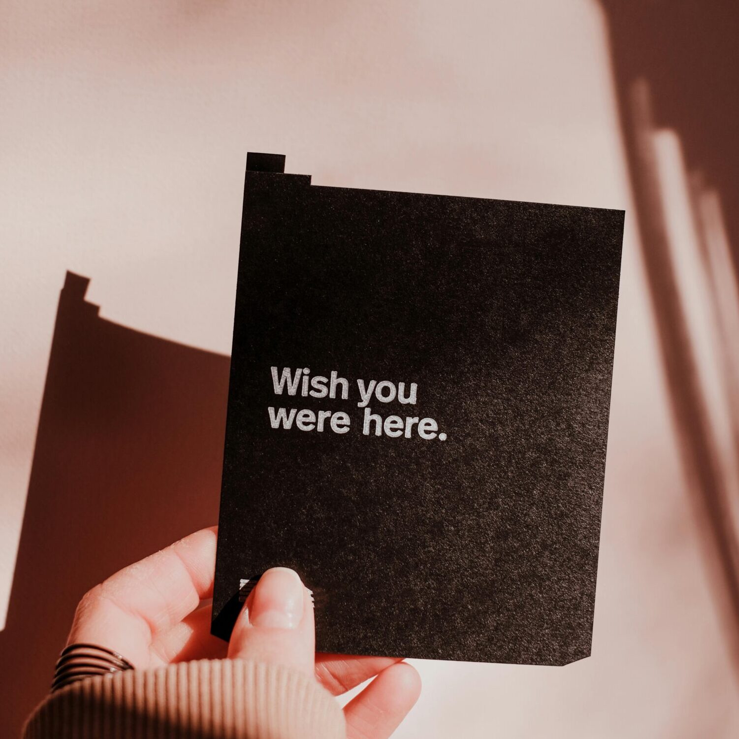 Minimalist photo of a hand holding a card with 'Wish you were here.' text and dramatic shadow play.